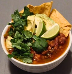 Bowl of beef chili with limes, cilantro, avocado, and chips.